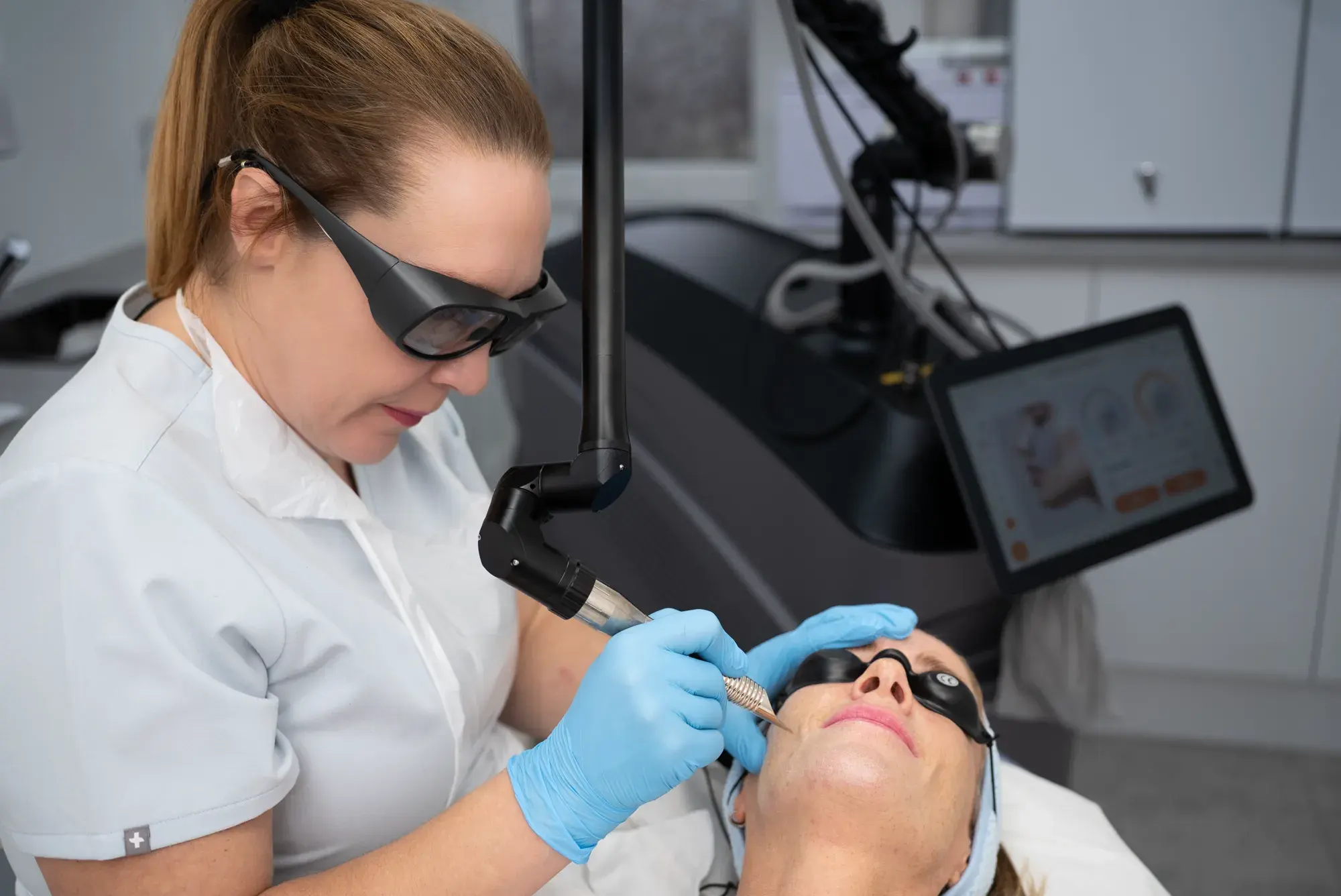 A practitioner in protective glasses treats a client's face with an erbium laser handpiece beside the machine