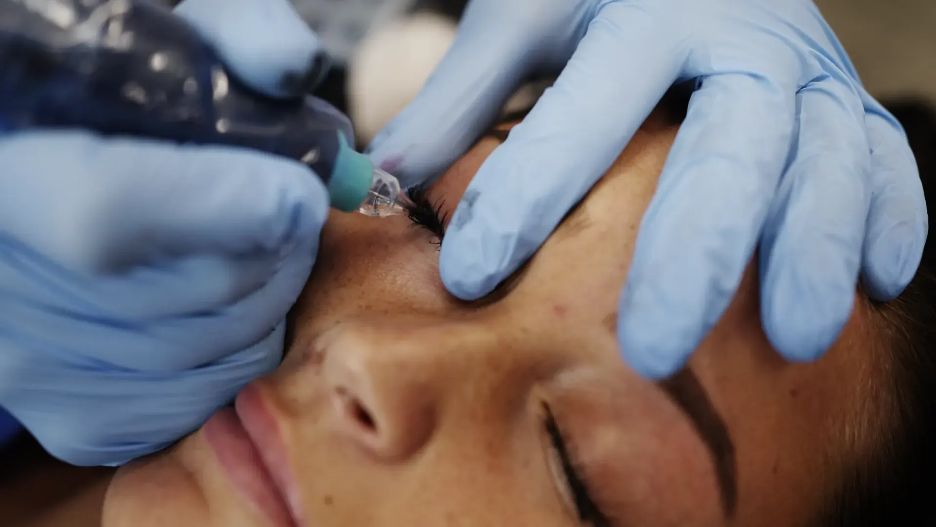 A practitioner holds a tattoo machine handpiece at a client's lash line
